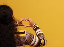 dekle pred rumenim zidom, ki z dlanmi ustvarja obliko srčka